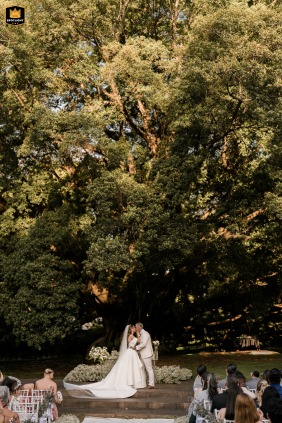   Caught in a loving kiss, the couple in São Paulo shares a moment filled with romance and happiness during their outdoor wedding ceremony under large trees.